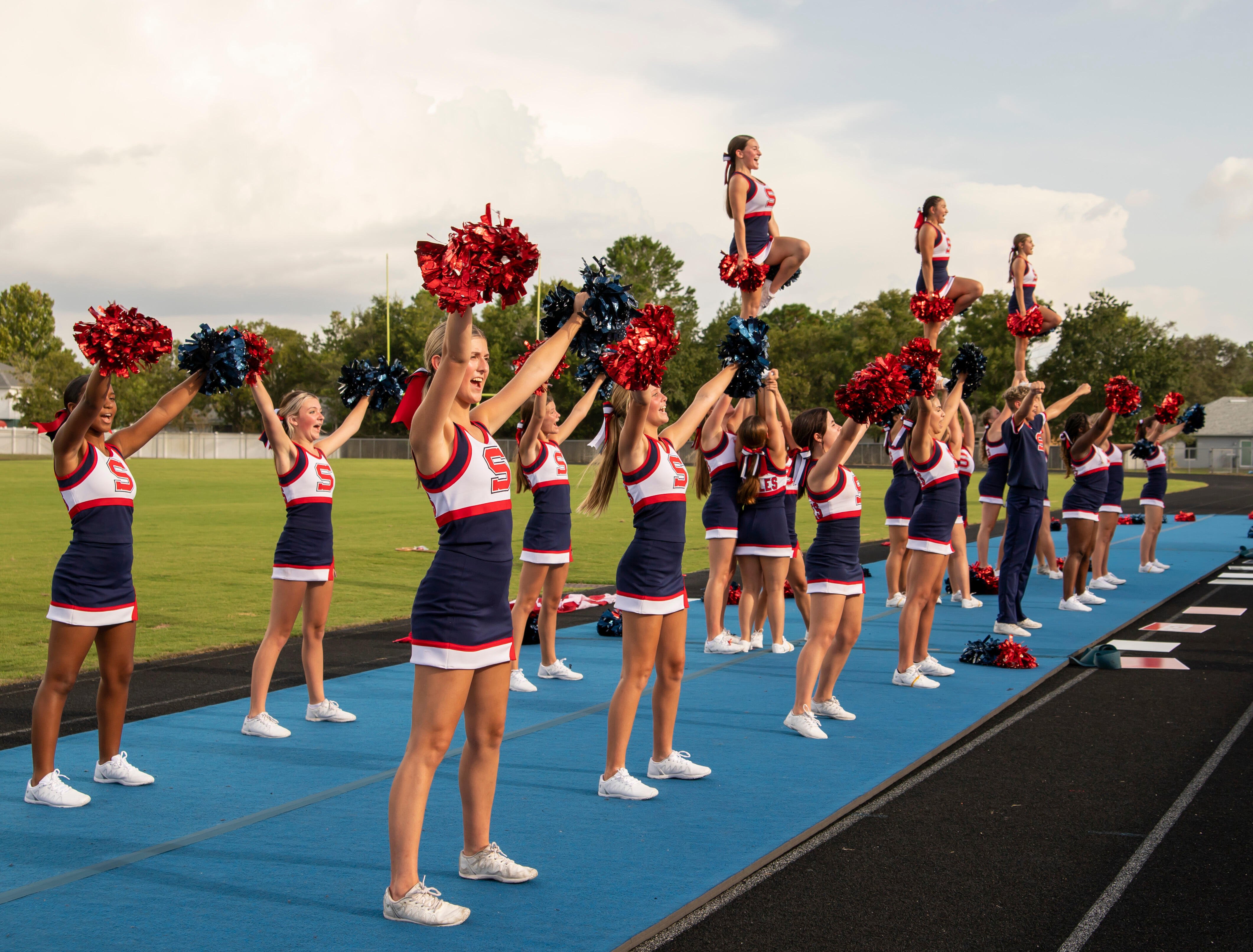 Un gruppo di cheerleader in divisa blu, rossa e bianca si esibisce su un tappeto azzurro a bordo campo. Alcune alzano i pon pon, altre sono sollevate in alto dai compagni. Sullo sfondo si vede un campo da sport all'aperto.
