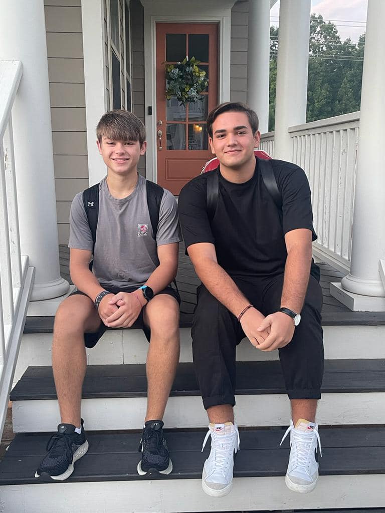 Due ragazzi sorridono seduti sui gradini di una veranda davanti a una casa. Entrambi indossano abiti casual e hanno uno zaino, come se fossero pronti per andare a scuola. L’atmosfera è rilassata e amichevole.
