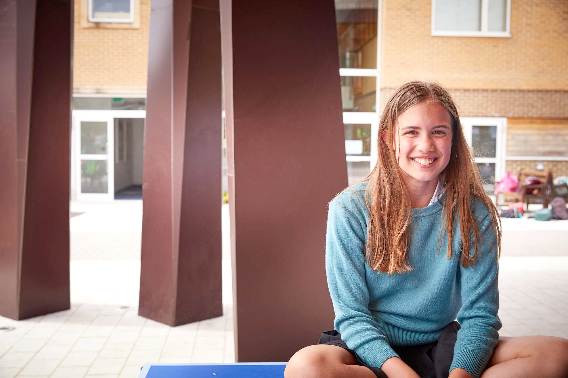Ragazza con capelli lunghi e maglione azzurro sorride seduta all’aperto, davanti a una struttura moderna con colonne marroni e un edificio scolastico sullo sfondo. L’atmosfera è serena e accogliente.