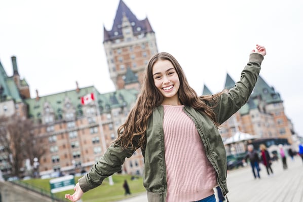 Una ragazza sorridente con le braccia aperte posa all’aperto davanti a un grande edificio storico con tetti verdi e una bandiera canadese. Indossa una giacca verde e un maglione rosa