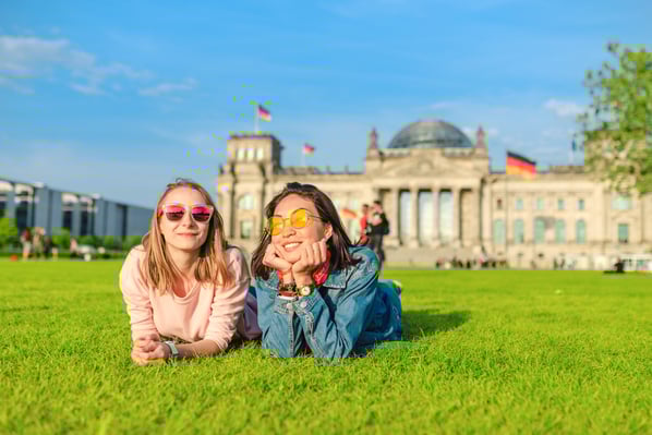 Due ragazze sdraiate su un prato verde sorridono con occhiali da sole, davanti al Reichstag di Berlino, in una giornata di sole.