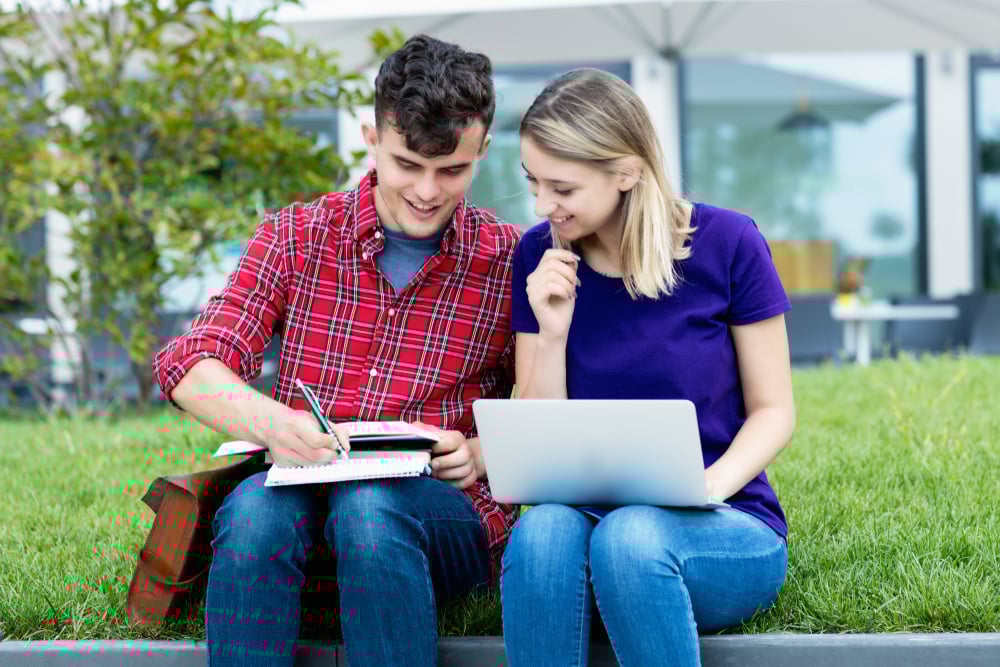 Due studenti seduti all’aperto sull’erba studiano insieme: uno scrive su un quaderno, l’altra usa un computer portatile. Entrambi sorridono.