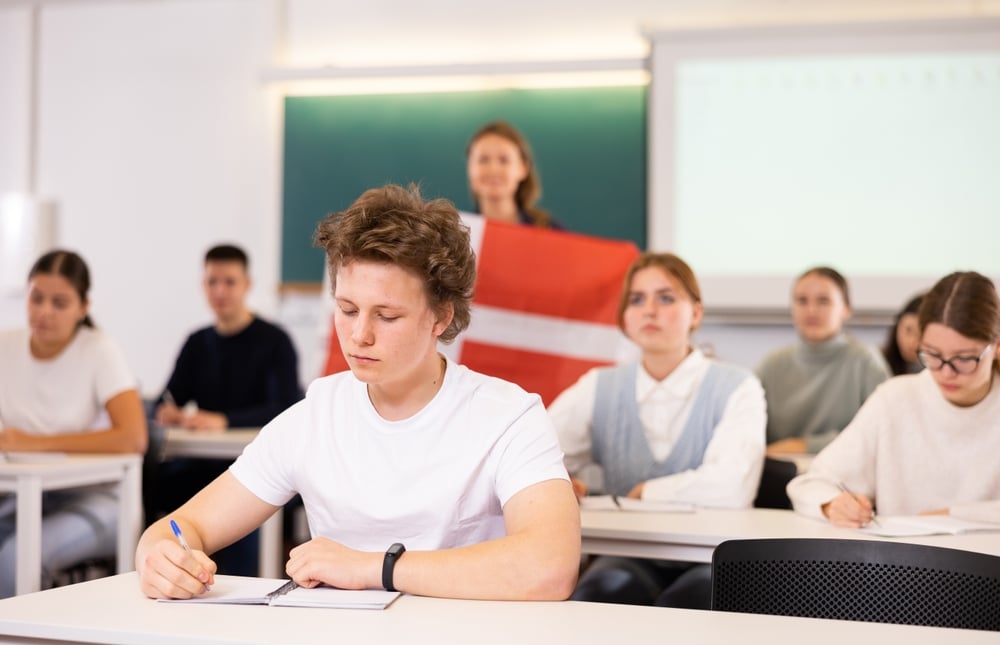 In un’aula scolastica, studenti seduti ai banchi scrivono e ascoltano. In primo piano un ragazzo biondo con maglietta bianca prende appunti. Sullo sfondo l’insegnante tiene una bandiera danese davanti alla lavagna.