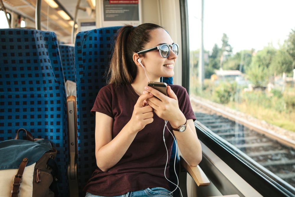 Ragazza con occhiali da sole e cuffiette ascolta musica e sorride guardando fuori dal finestrino di un treno in movimento.