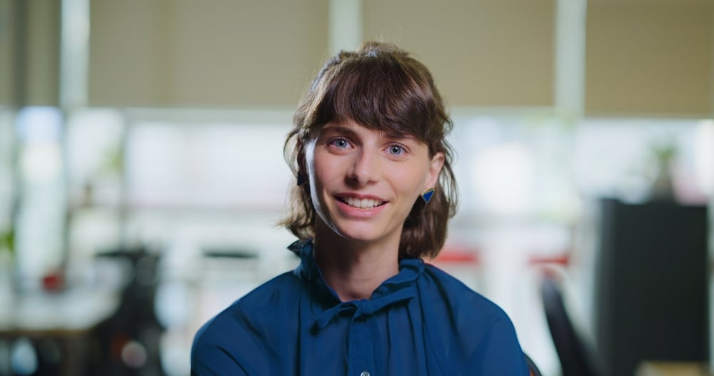 Ritratto di una giovane donna con capelli castani e occhi chiari che sorride, indossando una camicia blu. Lo sfondo è sfocato, in ambiente luminoso, probabilmente un ufficio o uno spazio di coworking.