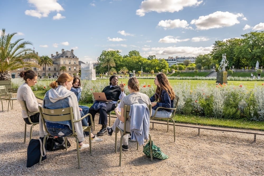 Sei ragazze siedono in cerchio su sedie da giardino, discutono e studiano insieme all’aperto in un parco fiorito, con edifici storici e statue sullo sfondo.