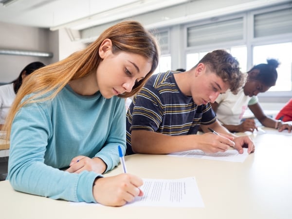 Studenti seduti in classe sono concentrati a scrivere durante un test o un compito in aula. In primo piano una ragazza con maglia azzurra.