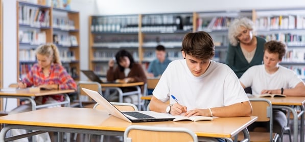 Studenti studiano e prendono appunti in una biblioteca. Un ragazzo in primo piano usa un computer portatile e scrive su un quaderno; un’insegnante cammina tra i banchi.