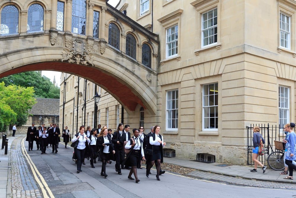 Un gruppo di studenti in uniforme scolastica cammina sotto un ponte storico ad arco tra edifici in pietra chiara. Alcuni studenti portano zaini e cartelle. Sul marciapiede si vedono anche alcune persone in abiti casual. L’atmosfera è vivace.