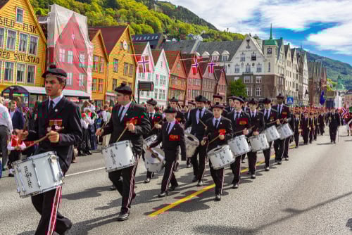 Una banda musicale in divisa sfila in parata su una strada, con ragazzi che suonano tamburi. Sullo sfondo, case colorate e bandiere norvegesi, davanti a una folla festosa.