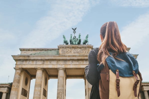 Una giovane con zaino osserva la Porta di Brandeburgo a Berlino vista da dietro, con il monumento e il cielo sullo sfondo.