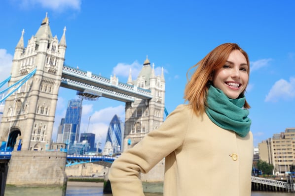 Una giovane donna con cappotto chiaro e sciarpa verde sorride davanti al Tower Bridge a Londra in una giornata soleggiata. Sullo sfondo si riconoscono anche i grattacieli della città.
