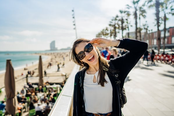 Una giovane donna con occhiali da sole sorride felice sulla passeggiata di una spiaggia affollata, con palme e il mare sullo sfondo in una giornata di sole.