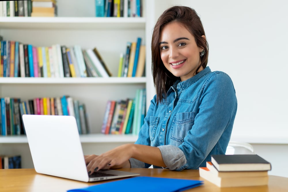 Una giovane donna sorride alla scrivania con un computer portatile, circondata da libri in una biblioteca o aula studio.