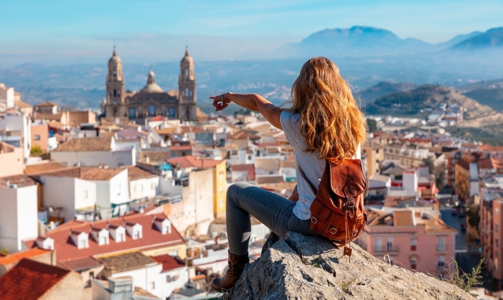 Una ragazza con zaino siede su una roccia e indica con il braccio verso una città e una cattedrale in lontananza, tra i tetti delle case e le montagne sullo sfondo.