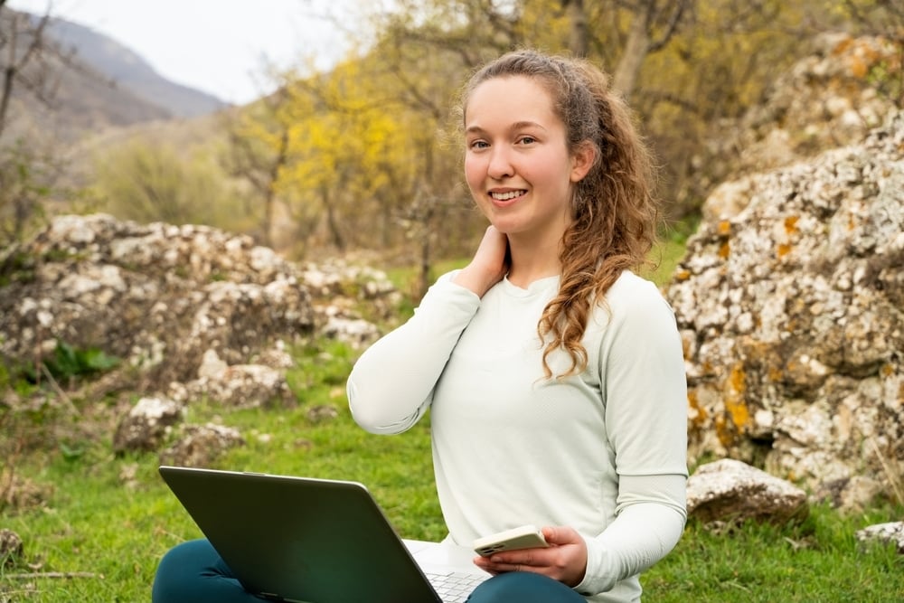 Una ragazza sorride seduta all’aperto su un prato tra rocce e alberi, con un computer portatile sulle gambe e uno smartphone in mano. Sembra studiare o lavorare nella natura.
