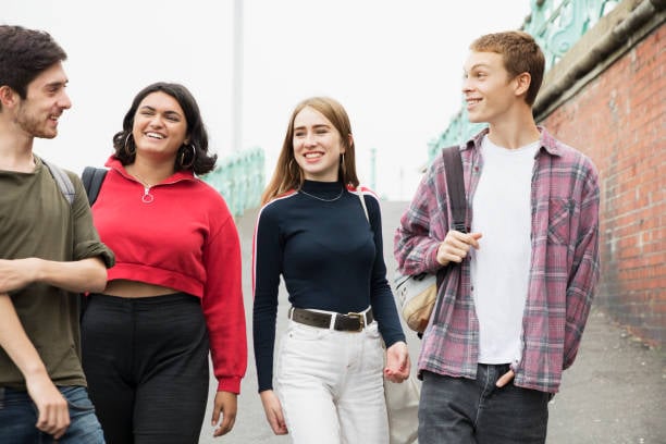 Quattro giovani camminano insieme all’aperto, sorridono e chiacchierano. Indossano abiti casual e zaini, sembrano studenti. Sullo sfondo c’è un muro di mattoni e una ringhiera verde. L’atmosfera è amichevole e rilassata.