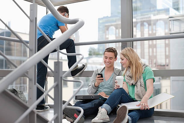 Due studenti sono seduti su delle scale interne vicino a grandi finestre; uno guarda il telefono, l’altra tiene una tazza e delle cartelle. Un terzo studente sta in piedi sulla scala. L’ambiente è luminoso e moderno, con vista sulla città.