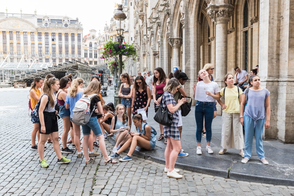Un gruppo di ragazze adolescenti si trova su una piazza di città storica, alcune sedute, altre in piedi vicino a un edificio antico con archi. L'atmosfera è di visita turistica. Sullo sfondo si vedono edifici storici e fiori appesi.