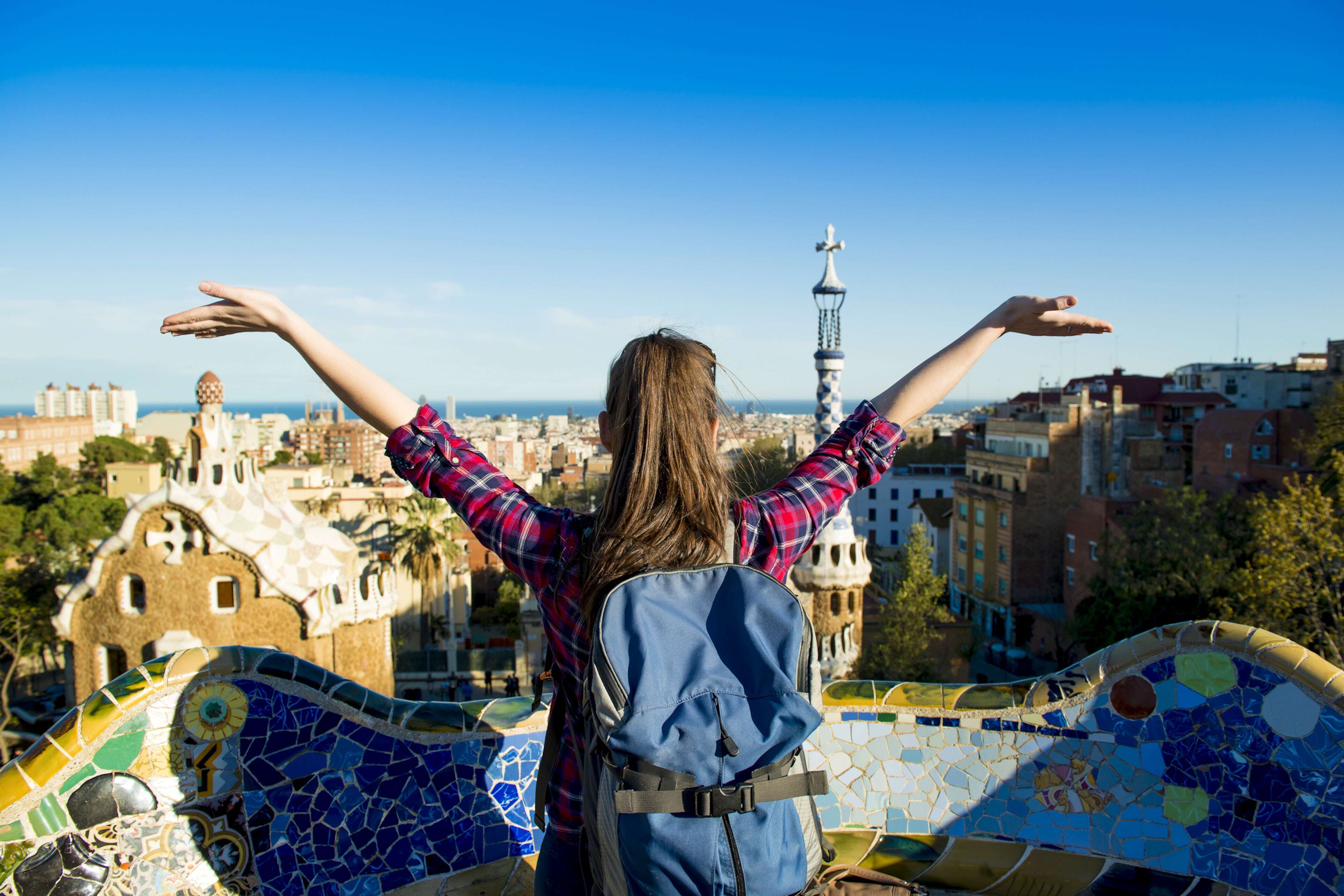 Ragazza con zaino e camicia a quadri, di spalle, a braccia aperte, ammira il panorama di una città colorata da un punto panoramico con mosaici. Sullo sfondo si vede il mare e l’architettura tipica di Barcellona.