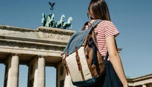 Ragazza di spalle con zaino, maglietta a righe rosse e capelli lunghi osserva la Porta di Brandeburgo a Berlino sotto un cielo azzurro. L’immagine trasmette un senso di viaggio e scoperta in una città europea.