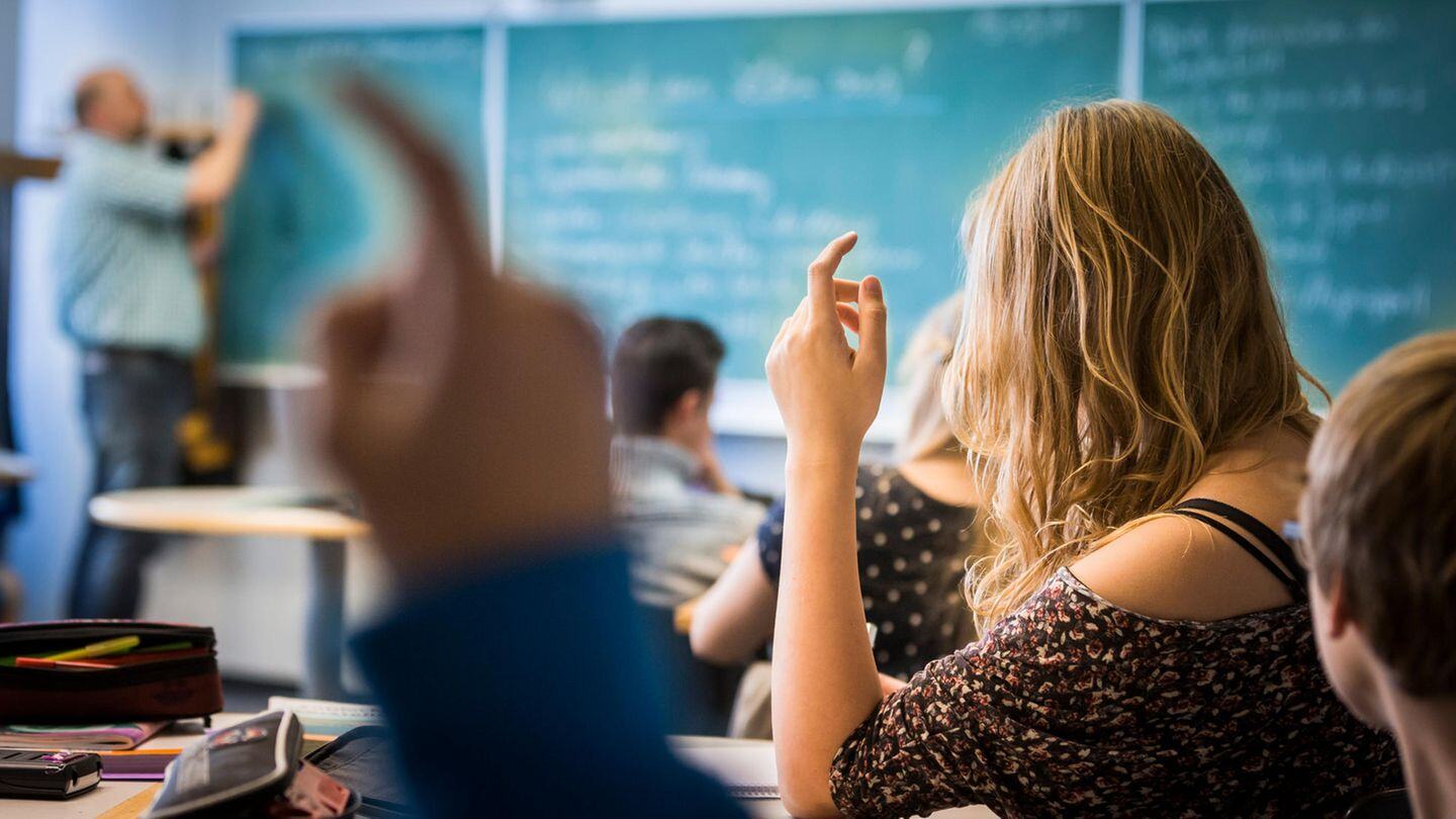 In un’aula scolastica, diversi studenti sono seduti ai banchi e alzano la mano per partecipare. In primo piano si vede una ragazza bionda di spalle. Sullo sfondo, un insegnante scrive alla lavagna.